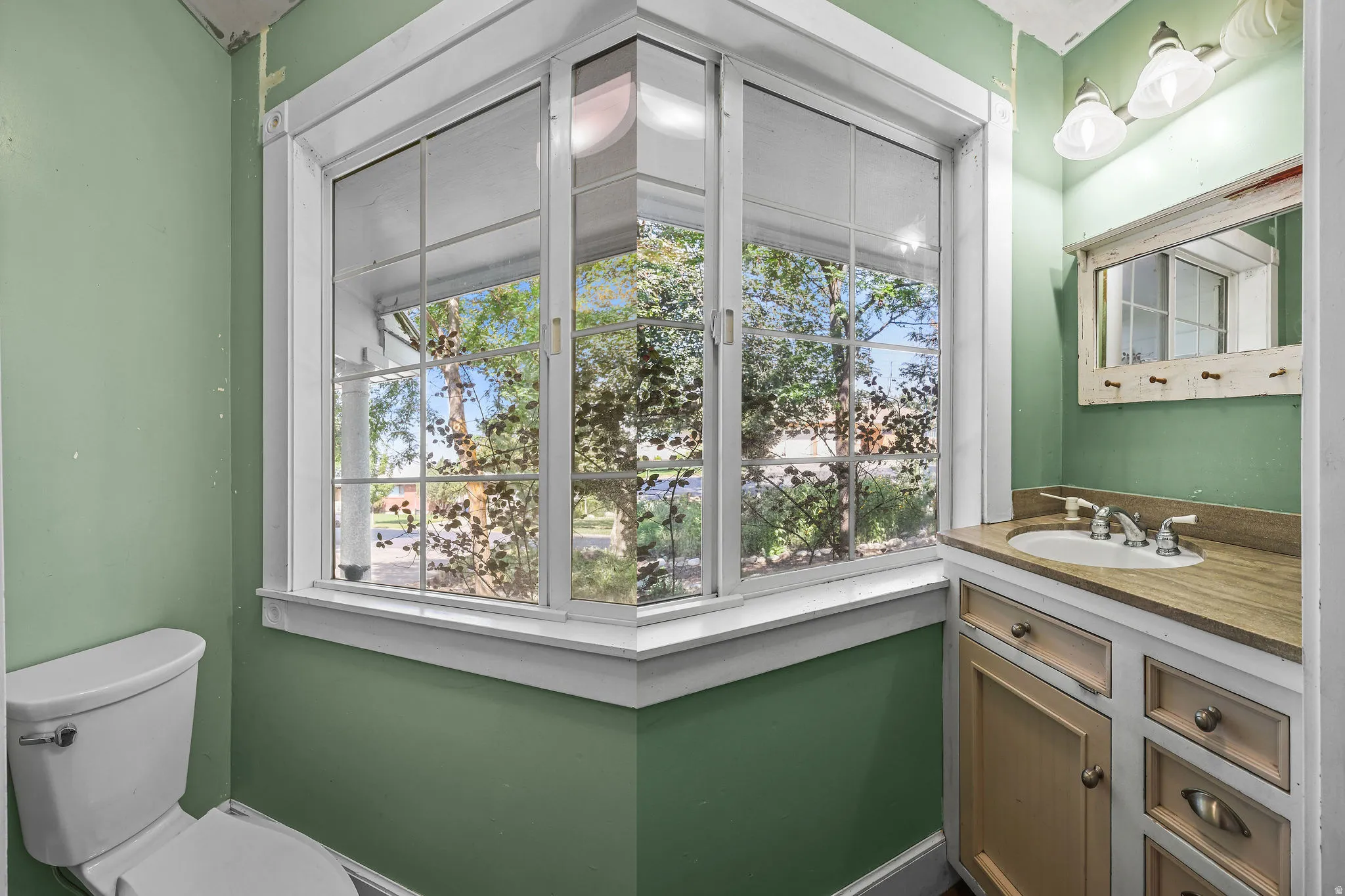 Bay window with white framing, built-in vanity with a single basin sink, wall-mounted light fixture, and a framed mirror