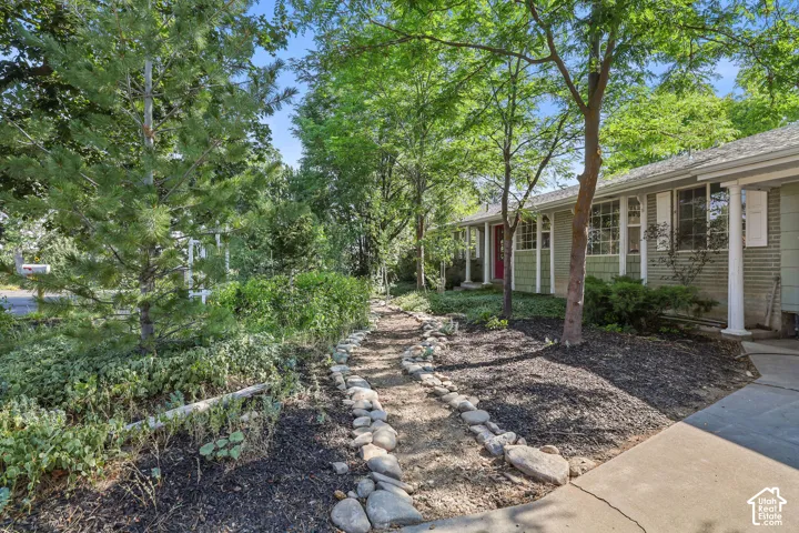 Exterior featuring green siding, white window shutters, mature trees, a stone-lined pathway, and a concrete driveway