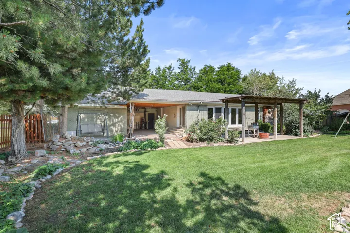 Expansive backyard with manicured lawn and mature tree coverage, anchored by a single-story residence featuring a covered patio, wood-beam pergola, and multi-panel sliding glass doors providing indoor-outdoor connectivity