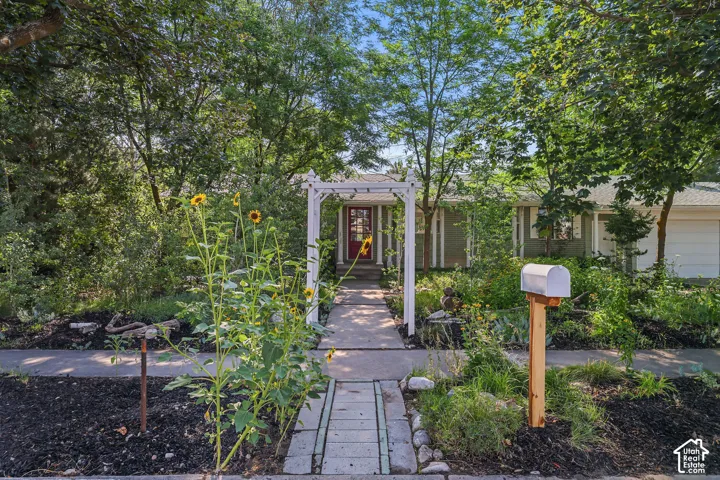 Front exterior featuring mature trees, a concrete pathway, a white arbor, a red entry door, and an attached garage