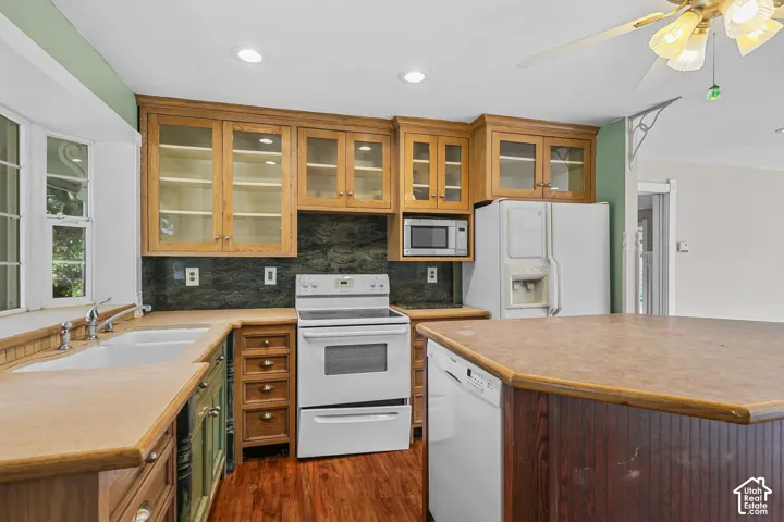 Wood-finish flooring, wood cabinetry with glass-front uppers, dual basin sink with gooseneck faucet, built-in microwave, and ceiling fan with integrated lighting