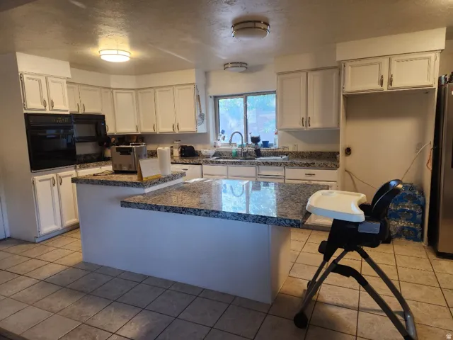 Spacious kitchen featuring white cabinetry with brushed nickel hardware, granite-topped island with seating, stainless steel appliances including built-in wall oven, tile flooring, recessed ceiling lighting, and a large window overlooking the landscape