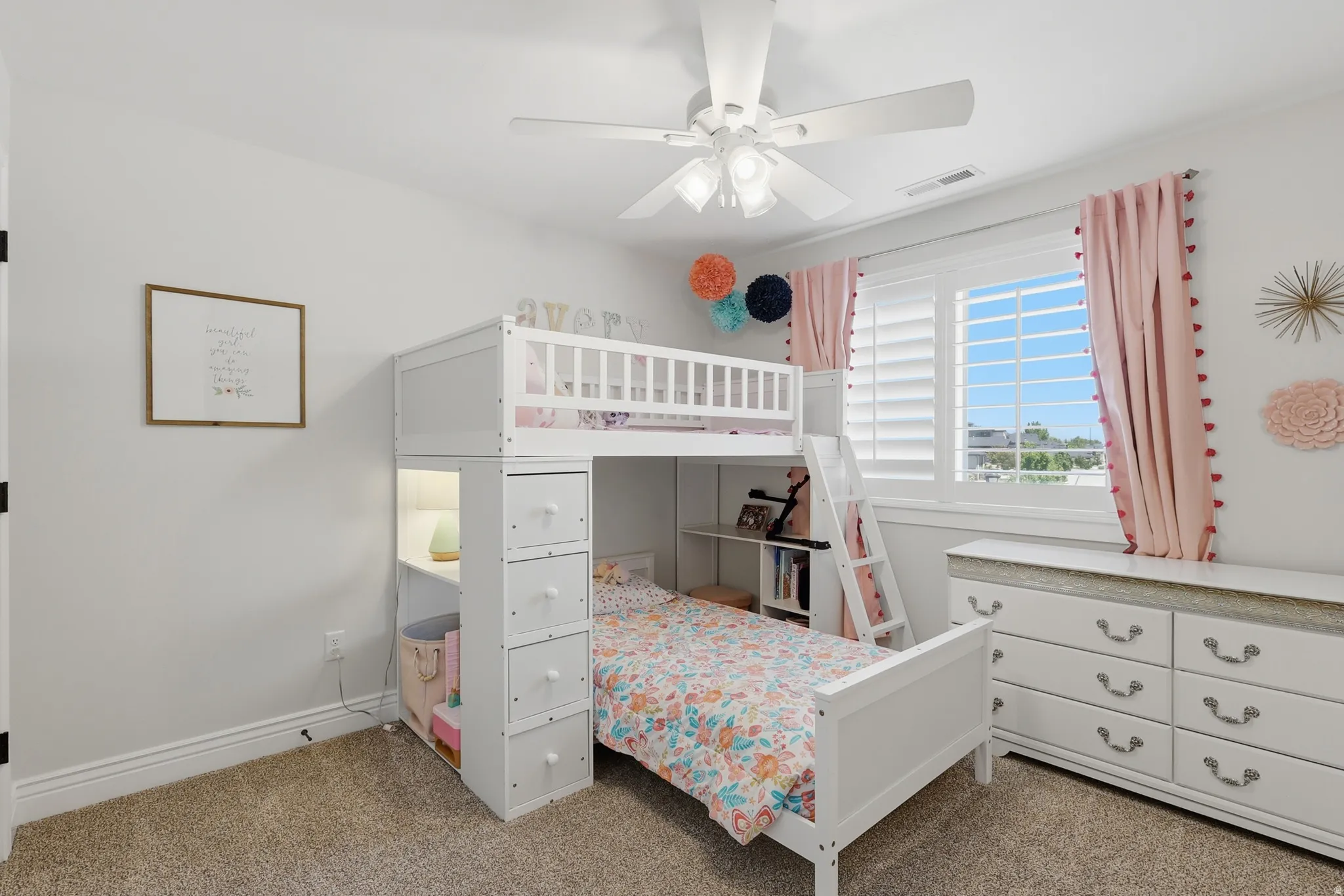White bunk bed with built-in storage, light-colored walls, ceiling fan with integrated lighting, window with white plantation shutters, and carpeted flooring