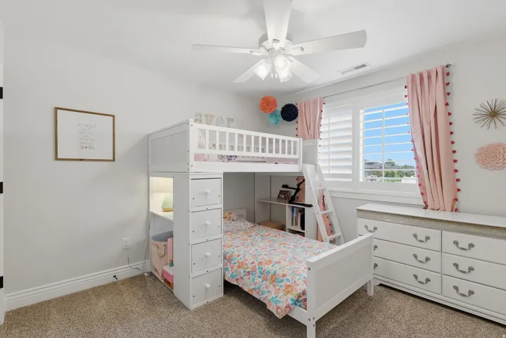 White bunk bed with built-in storage, light-colored walls, ceiling fan with integrated lighting, window with white plantation shutters, and carpeted flooring