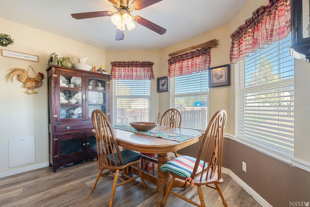 Bright dining area featuring a wood-finish table with coordinating chairs, abundant natural light from multiple windows with red plaid valances and shutters, ceiling fan with integrated lighting, and a wood-finish china cabinet with glass doors and lower