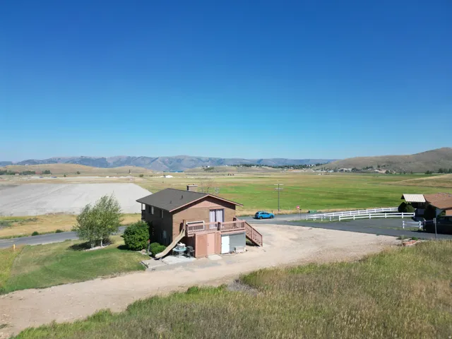 Rustic ranch compound situated on expansive grounds featuring a brown metal-sided residence with covered deck entry, detached outbuildings, gravel driveway, white fencing, and pastoral acreage with mountain backdrop