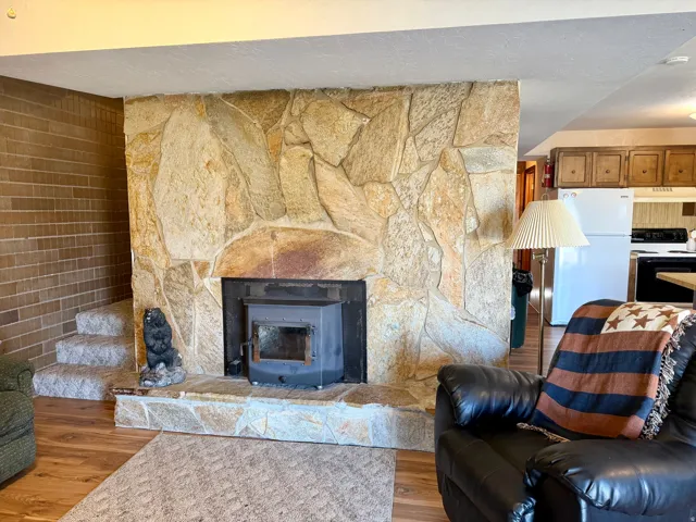 Stone fireplace with wood-burning stove insert, hardwood flooring throughout the living area, leather seating, built-in cabinetry visible in the adjoining kitchen, and warm ambient lighting from recessed ceiling fixtures