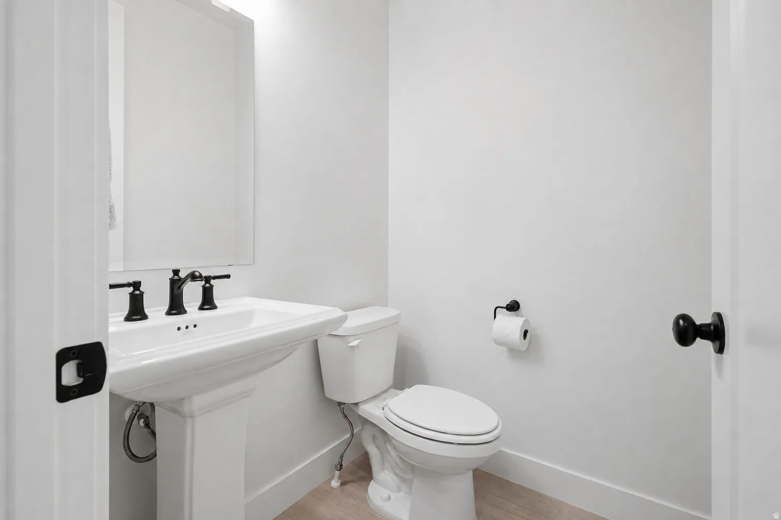 Pedestal sink with black matte faucet, rectangular mirror, toilet, and light wood-finish flooring