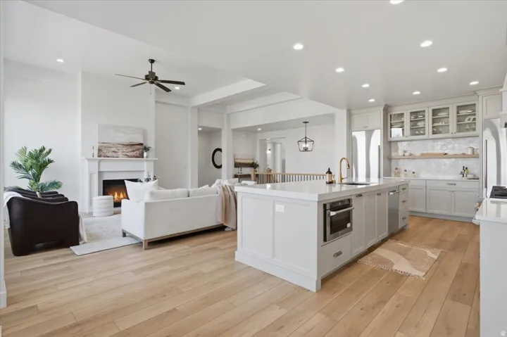 Open-concept living space featuring wood-finish flooring, a kitchen island with built-in appliances, white shaker cabinetry with glass inserts, and a fireplace with a mantel