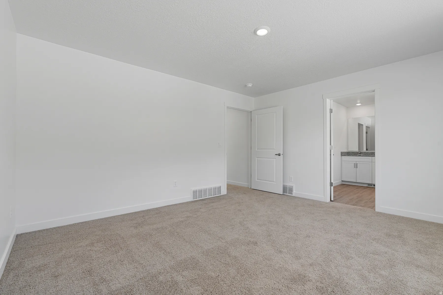Spacious carpeted bedroom with neutral wall finishes, recessed ceiling lighting, and direct access to an ensuite bathroom and hallway entry
