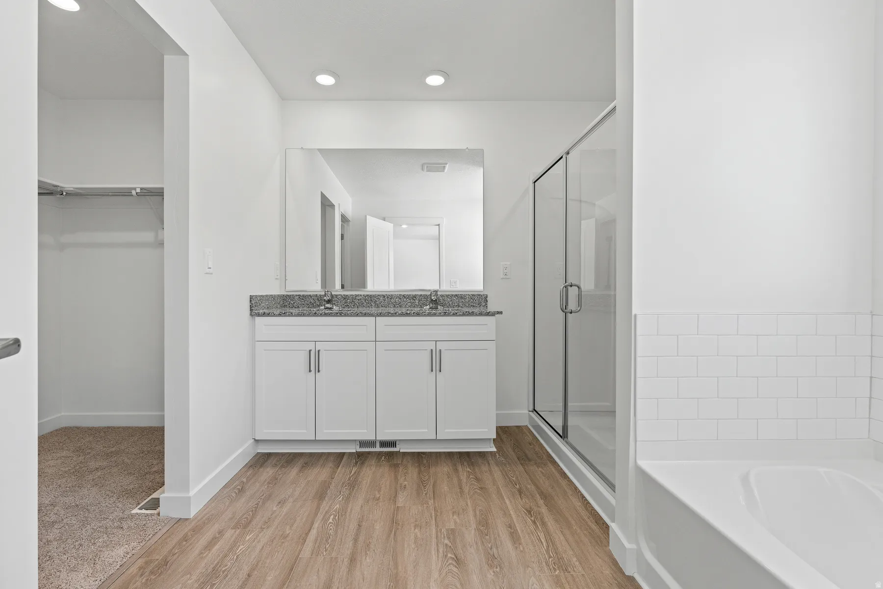 Spa-inspired primary bathroom featuring dual vanity with white cabinetry and brushed nickel hardware, gray tile backsplash, frameless glass walk-in shower enclosure, and wood-finish flooring throughout