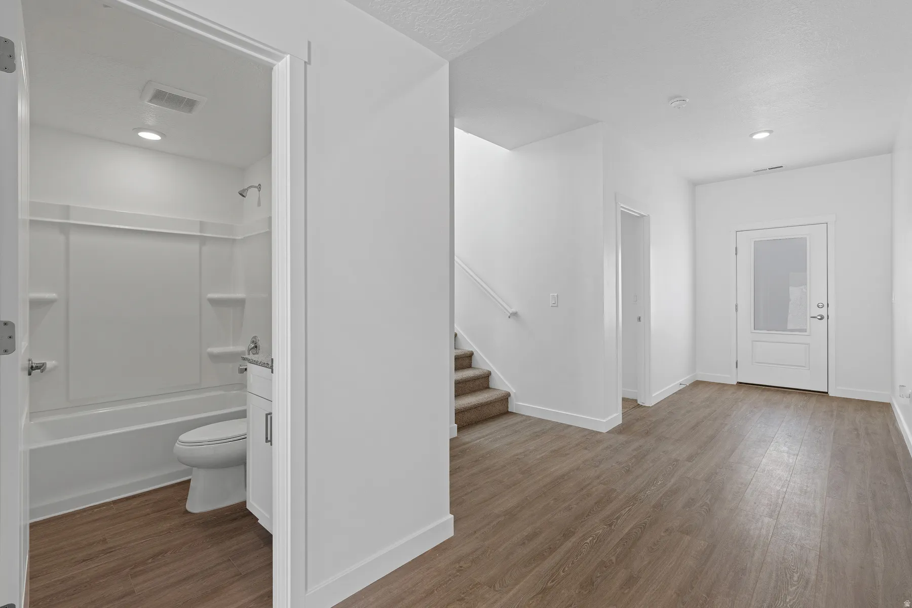 Bright, open basement space featuring wood-finish flooring throughout, a full bathroom with white subway tile surround and modern fixtures, and a staircase with wood treads