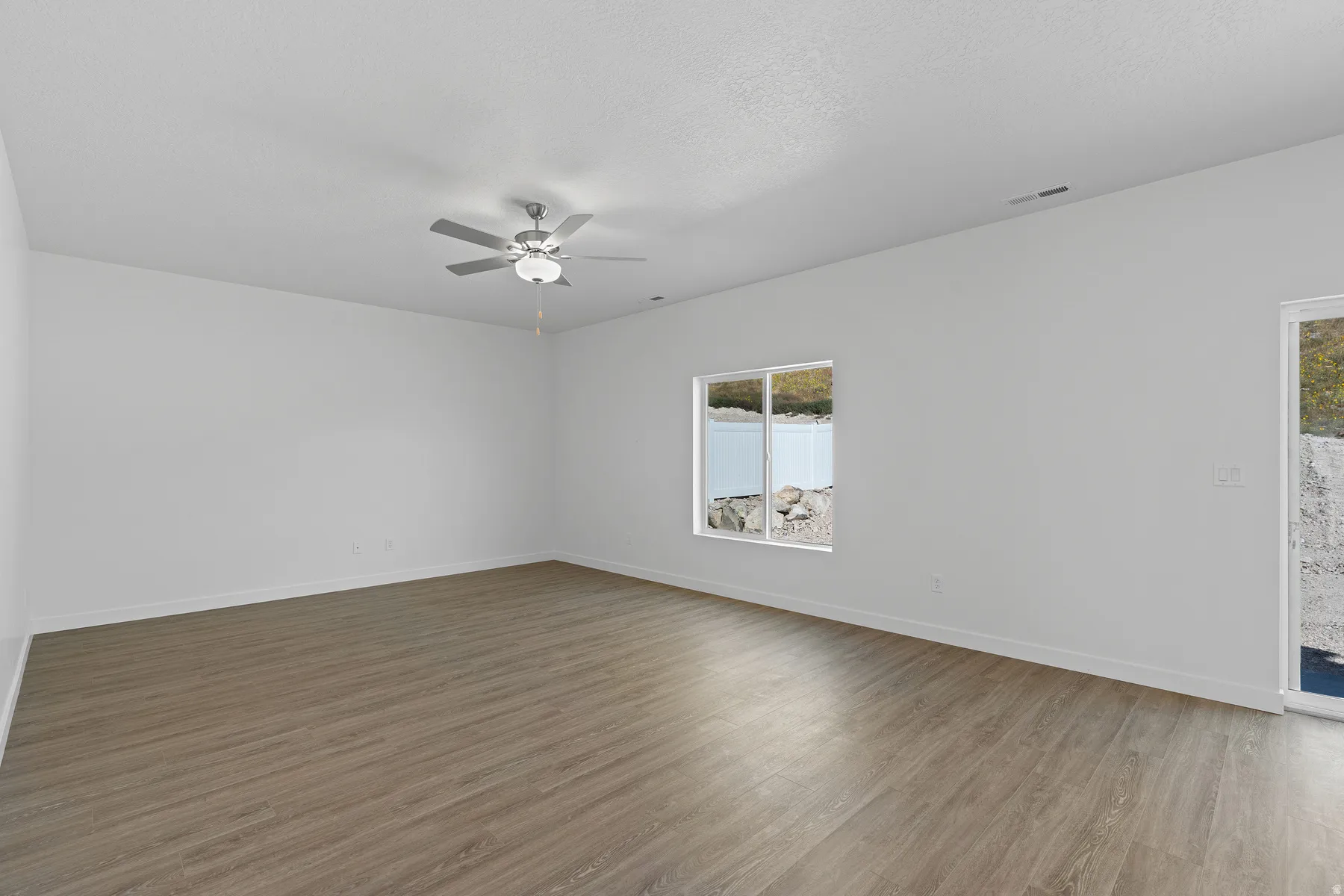 Spacious room featuring wood-finish flooring, white walls, a ceiling fan with integrated lighting, and a large window with a water and landscape backdrop