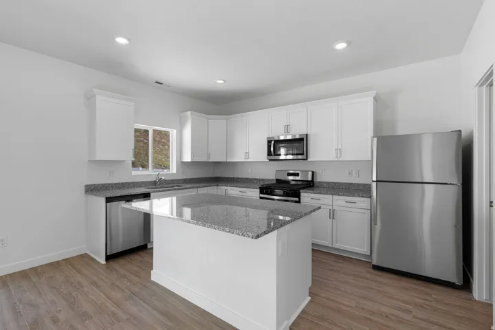 Modern kitchen featuring white cabinetry, stainless steel appliances including a built-in microwave and refrigerator, gray quartz countertops, a centered island with additional counter space, wood-finish flooring, and recessed ceiling lighting