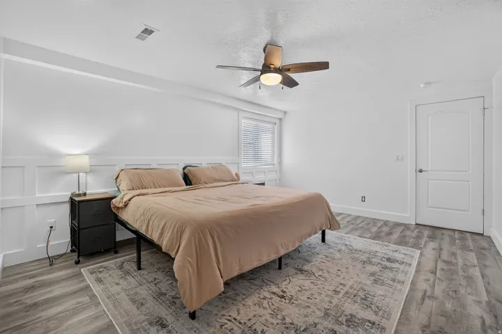 Wood-finish flooring, ceiling fan with integrated lighting, window with blinds, interior door with polished hardware, and wainscoting detail