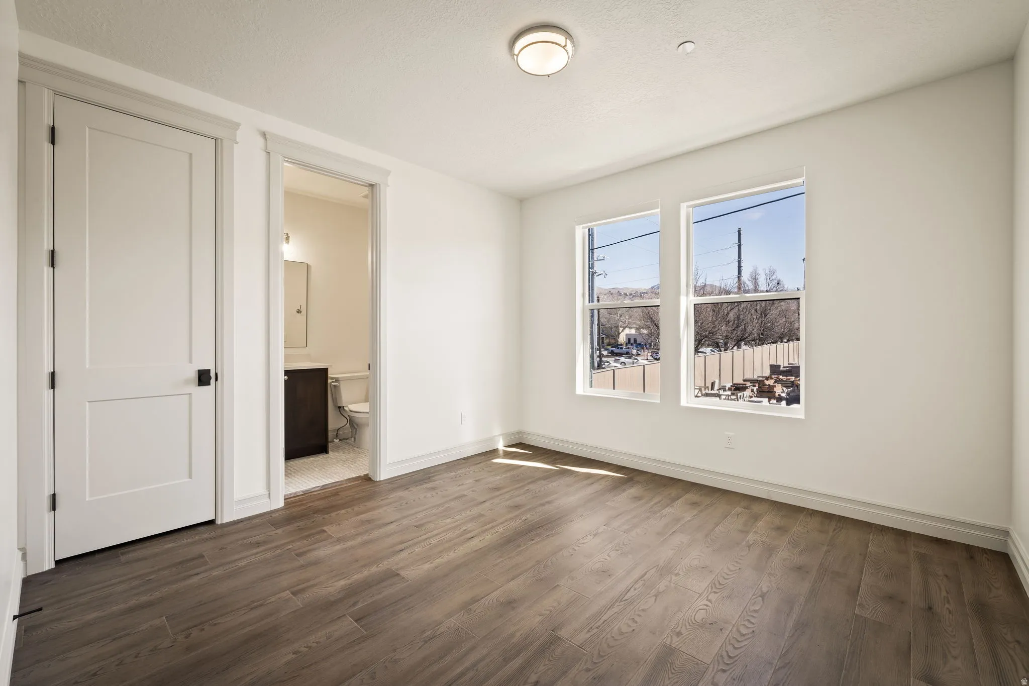 Bright corner bedroom showcasing hardwood flooring, dual windows with natural light, white six-panel closet doors, and an en-suite bathroom with direct access