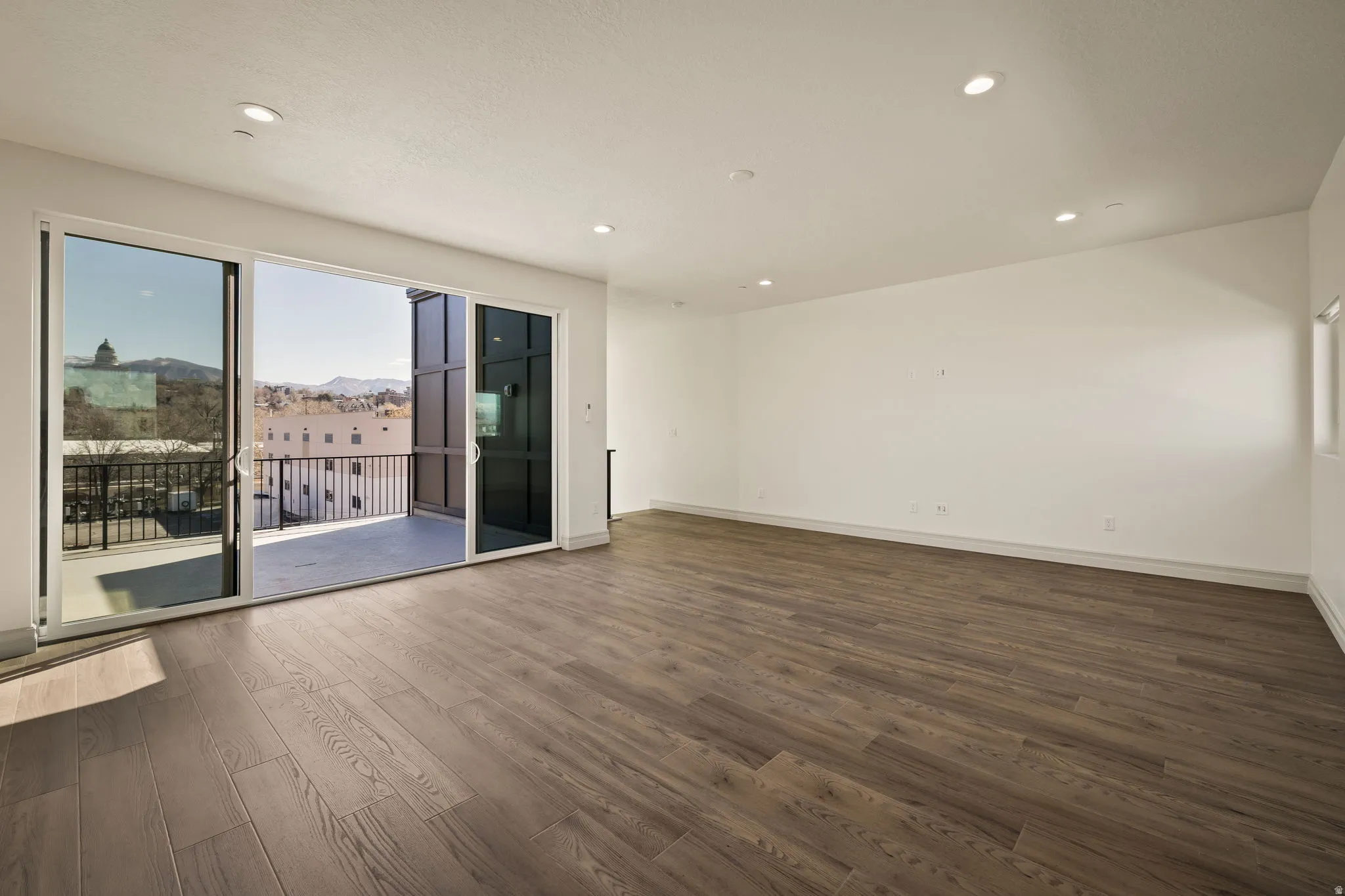 Spacious corner room featuring premium wood-finish flooring, recessed ceiling lighting, and expansive glass sliding doors with direct access to a private balcony overlooking the surrounding landscape