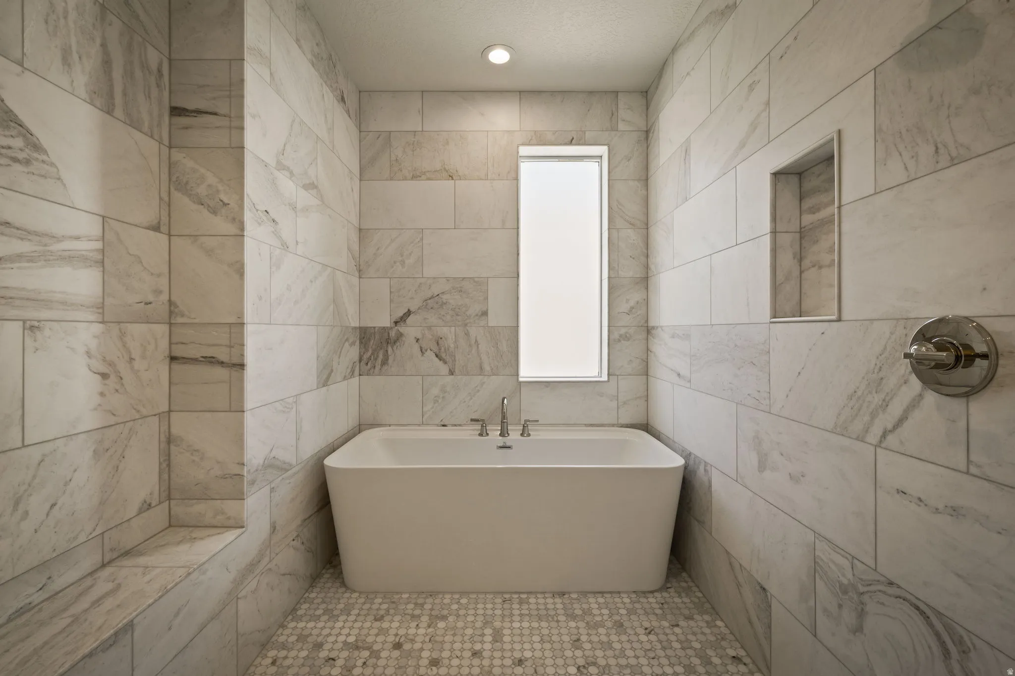 Freestanding soaking tub, tiled walls with integrated niche, built-in shower bench, and mosaic tile flooring
