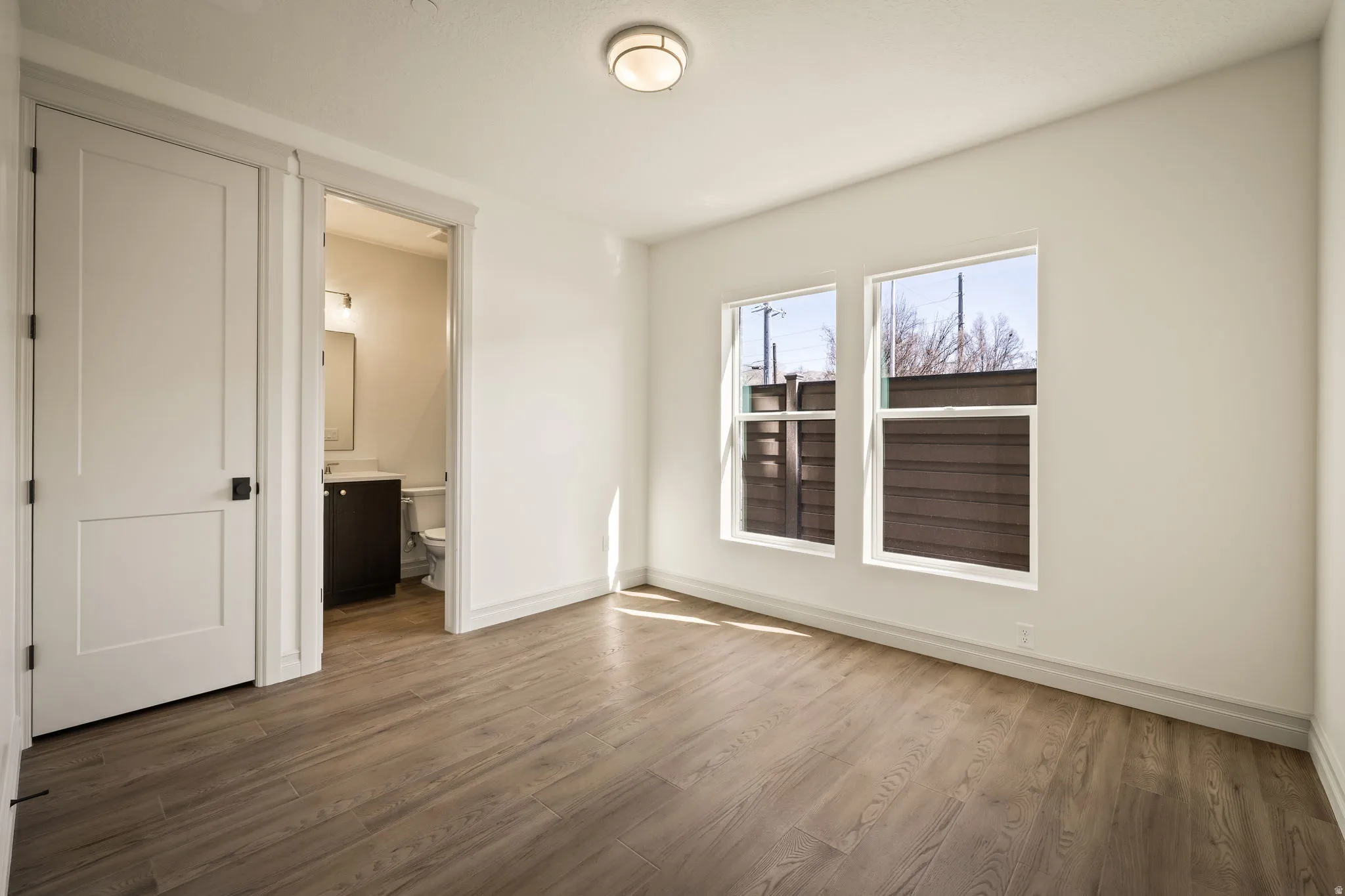 Bright corner bedroom with wood-finish flooring, dual windows with cellular shades, and direct access to a full bathroom