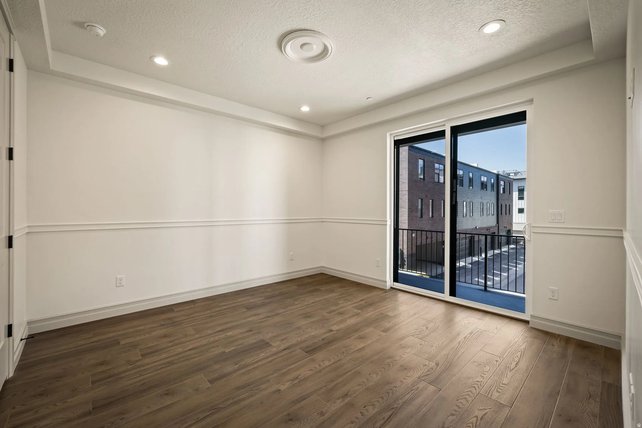 Bright, light-filled room featuring pristine wood-finish flooring, crisp white wall paneling with horizontal trim detail, recessed ceiling lighting, and dual glass sliding doors opening to an exterior balcony with metal railing