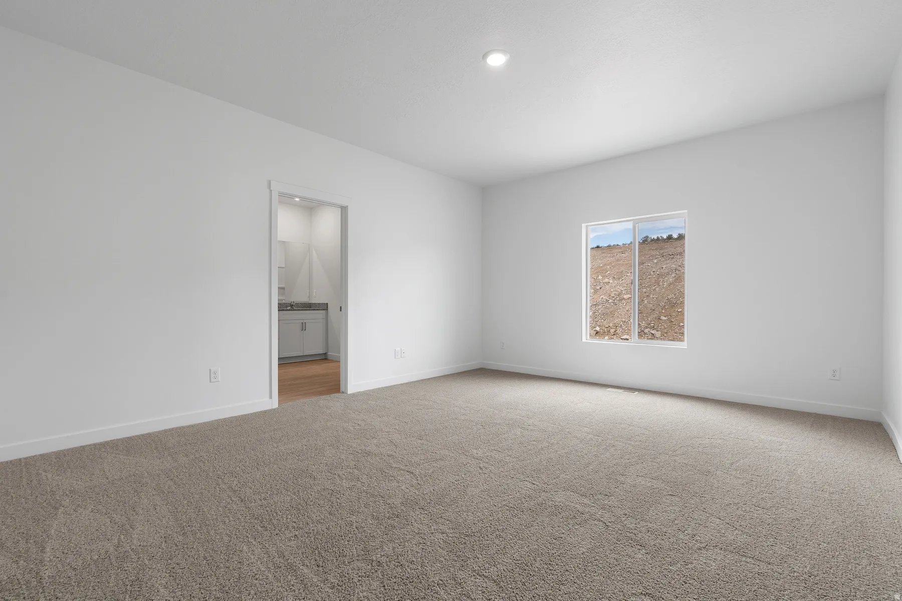 Spacious bedroom showcasing neutral carpeting, abundant natural light from a large window, recessed ceiling lighting, and direct access to an ensuite bathroom with glass-enclosed shower