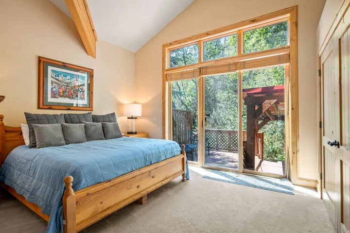 Primary bedroom suite flooded with natural light through expansive glass doors and transom windows, featuring solid wood bed frame, neutral carpet flooring, recessed ceiling detail, and direct access to outdoor deck and landscaped grounds