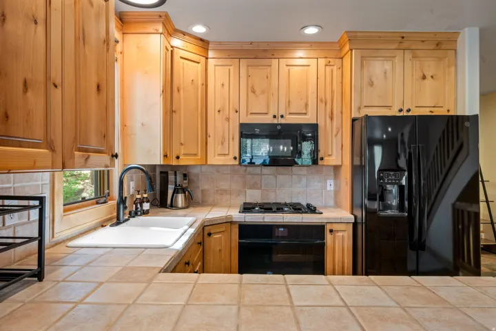 Warm wood cabinetry with raised-panel doors and brushed hardware frames this kitchen, featuring tile countertops, a gas cooktop with matching backsplash, and stainless steel appliances including a side-by-side refrigerator and built-in microwave