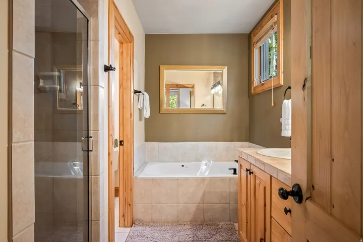 Spa-inspired primary bathroom featuring a frameless glass-enclosed shower with tile surround, a soaking tub with tile apron, dual vanity with wood cabinetry and black hardware, a framed mirror, and warm neutral wall finishes