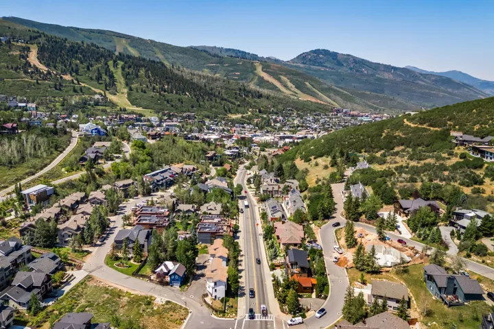 Aerial view showcasing a mountain community nestled within evergreen-forested slopes, featuring tree-lined residential streets, established landscaping throughout the neighborhood, and distant mountain peaks creating a scenic backdrop