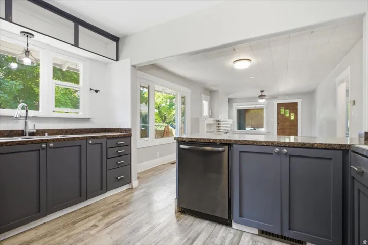 Modern kitchen featuring dark cabinetry with contrasting wood-finish flooring, stainless steel appliances including a built-in dishwasher, granite countertops, and abundant natural light from multiple windows and glass doors opening to outdoor space