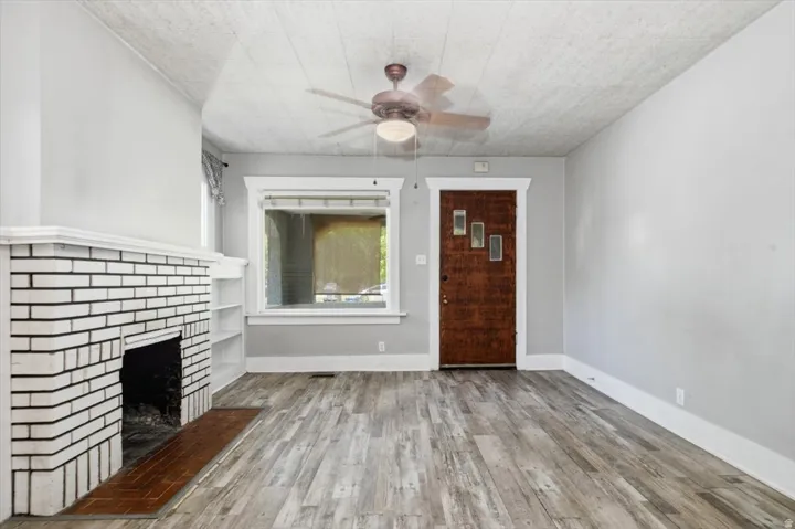 Bright living space featuring a wood-finish plank floor, white subway tile fireplace with dark hearth, ceiling fan with integrated lighting, and large window with white trim