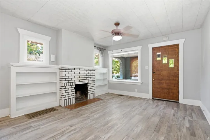 Bright open-concept living space featuring hardwood-finish flooring, a brick fireplace with white surround and built-in shelving, ceiling fan with integrated lighting, large window with natural light, and a solid wood entry door