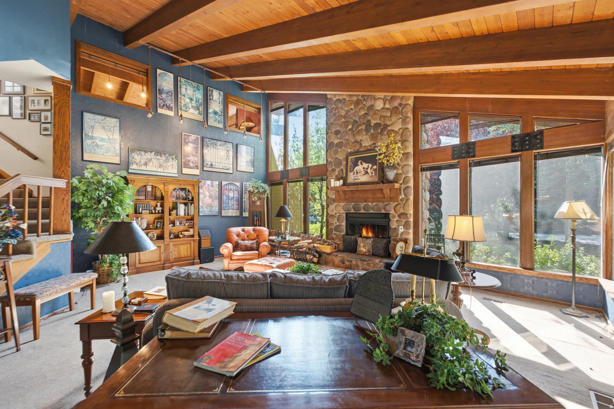 Vaulted wood-paneled ceiling with exposed beams, stone-clad fireplace, built-in wooden shelving, and large picture windows