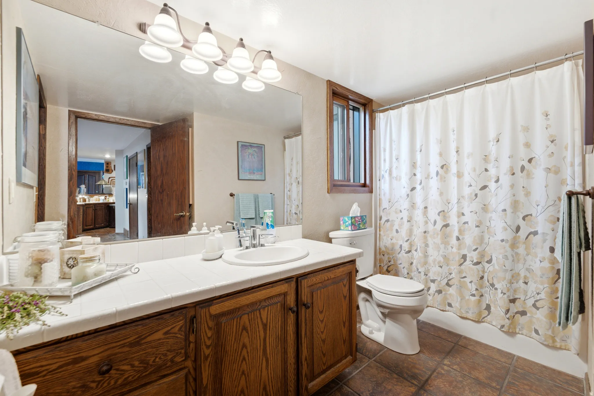 Tile flooring, wood-finish vanity with a single basin sink, white tiled countertop, wall-mounted light fixture, and an operable window