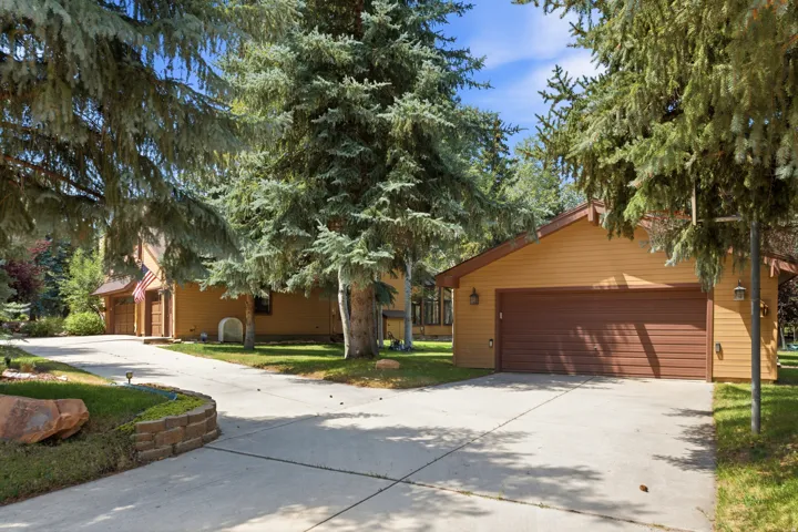 Detached garage on addional lot.  Expansive concrete driveway, attached garage with brown panel door, exterior wall sconces, and surrounding mature evergreen trees
