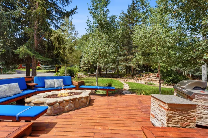 Expansive wood-decked patio featuring a stone-clad fire pit, built-in outdoor kitchen with stone veneer, and mature trees