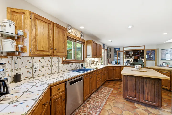 Wood cabinetry with glass shelving, tile countertops and backsplash, stainless steel dishwasher, and a wood-top kitchen island