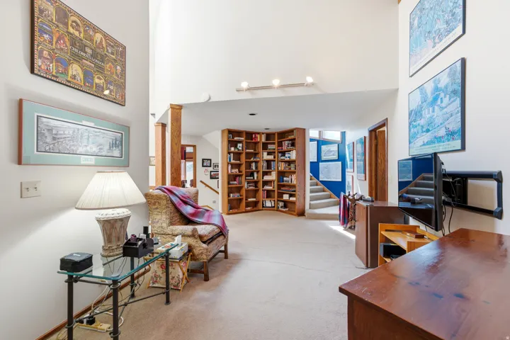 Vaulted ceiling, built-in shelving, wood-finish columns, carpeted flooring, and a staircase with a wood handrail