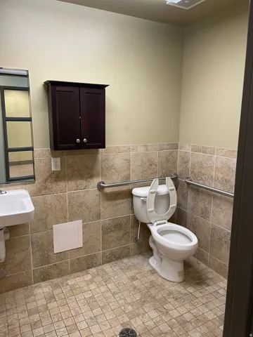 Wall-mounted vanity cabinet, ceramic tile wainscoting, white toilet, wall-mounted grab bars, and mosaic tile flooring