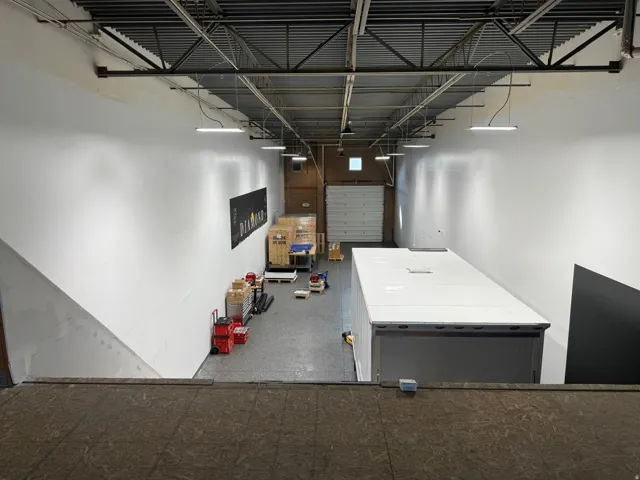Industrial interior with high ceilings, exposed structural beams, suspended linear lighting, a roll-up door, and polished concrete flooring