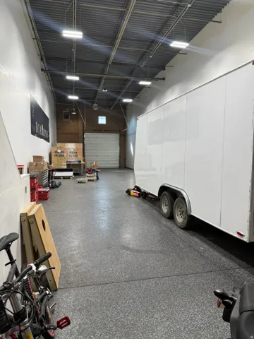 Spacious garage featuring epoxy-coated concrete flooring, high ceilings with exposed structural beams, multiple LED ceiling fixtures, and ample storage space with built-in wall cabinetry