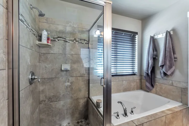 Spa-inspired primary bathroom featuring a frameless glass-enclosed walk-in shower with tile surround and rainfall showerhead, freestanding soaking tub with deck-mounted faucet, and natural light from a horizontal-slat window