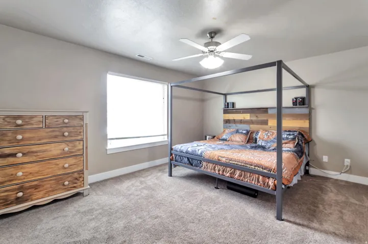 Spacious primary bedroom featuring a metal canopy bed frame, wood-finish dresser with multiple drawers, neutral carpeting, ceiling fan with integrated lighting, and large picture window allowing natural light throughout the room