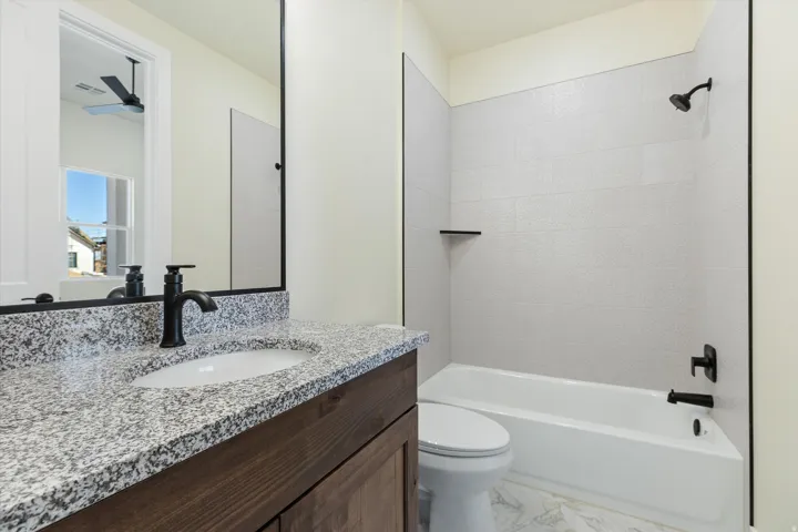 Modern bathroom featuring a speckled stone countertop with an undermount sink, matte black faucet and hardware, wood-finish vanity cabinetry, frameless glass shower enclosure, and combined tub-shower with subway tile surround and rainfall showerhead