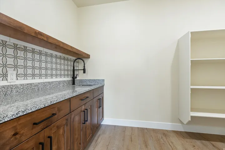 Wood-finish flooring, granite countertop, textured tile backsplash, dark-finished cabinetry, and a black-finish sink faucet