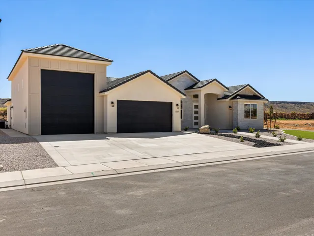 Modern residential compound featuring a striking two-tone exterior with cream and charcoal accents, multiple garage bays with black overhead doors, clean concrete driveway, and contemporary architectural detailing including peaked rooflines and geometric