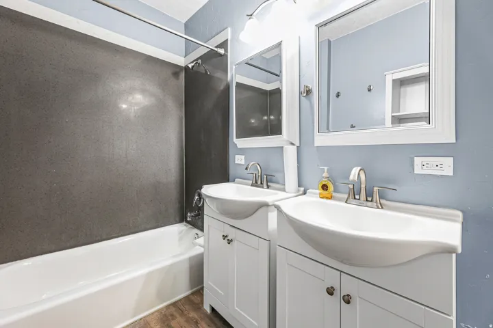 Dual vanities with white cabinetry, a built-in shower/tub combination, gray shower surround, and wood-finish flooring