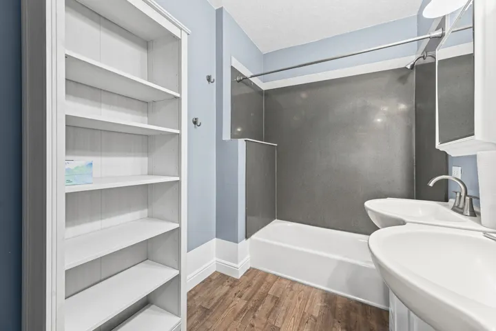 Wood-finish flooring, white built-in shelving, white bathtub with dark gray surround, dual pedestal sinks with brushed nickel faucets, and a recessed medicine cabinet