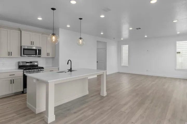 Modern open-concept kitchen and living space featuring white shaker-style cabinetry, stainless steel appliances, quartz countertops, a large center island with waterfall edge, pendant lighting, wood-finish flooring throughout, recessed ceiling lights, and