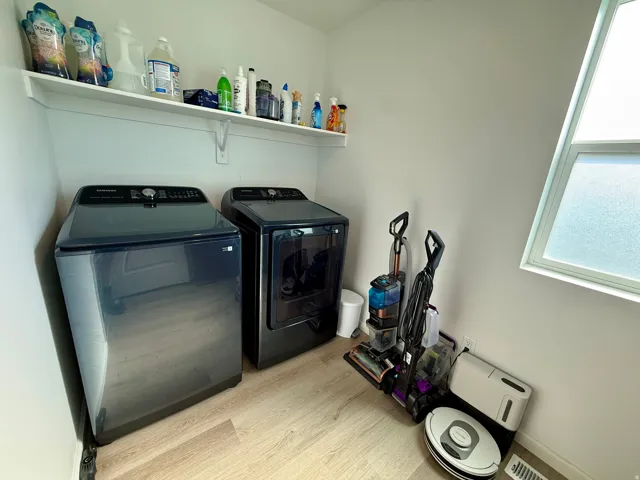 Modern laundry room featuring side-by-side washer and dryer with black stainless steel finishes, wood-finish flooring, ample shelving for storage and supplies, and abundant natural light from a large window